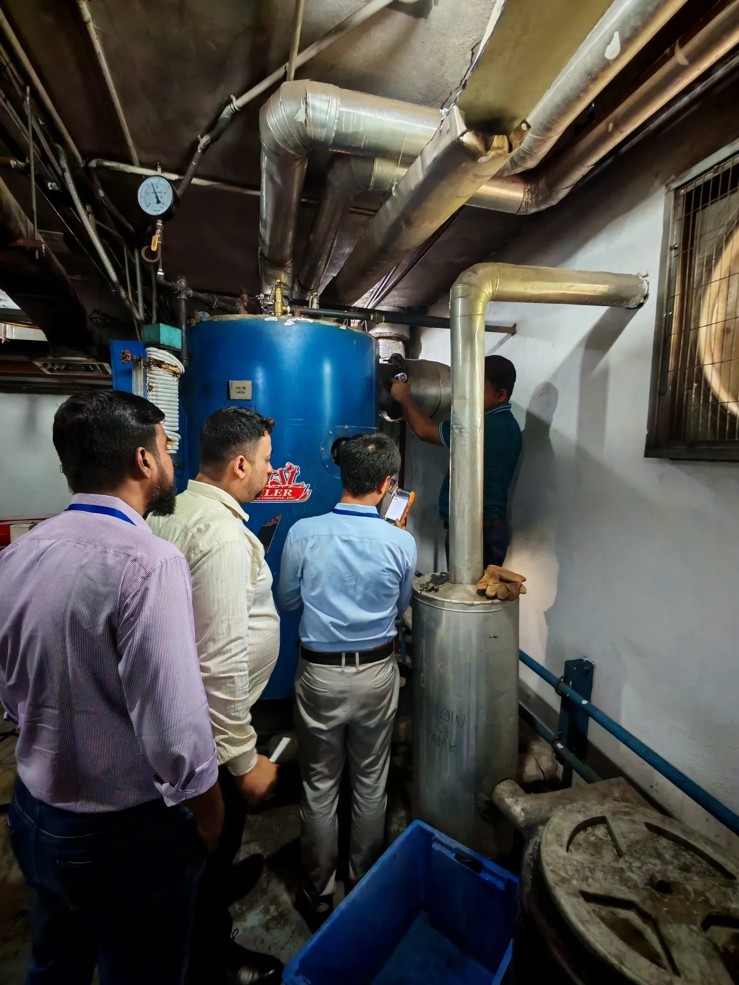 Auditors gathered around a boiler and chemical dosing tank in a plant room